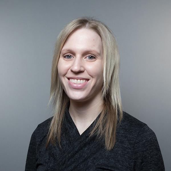 Professional portrait of a woman wearing black with blonde hair, against a gray background.